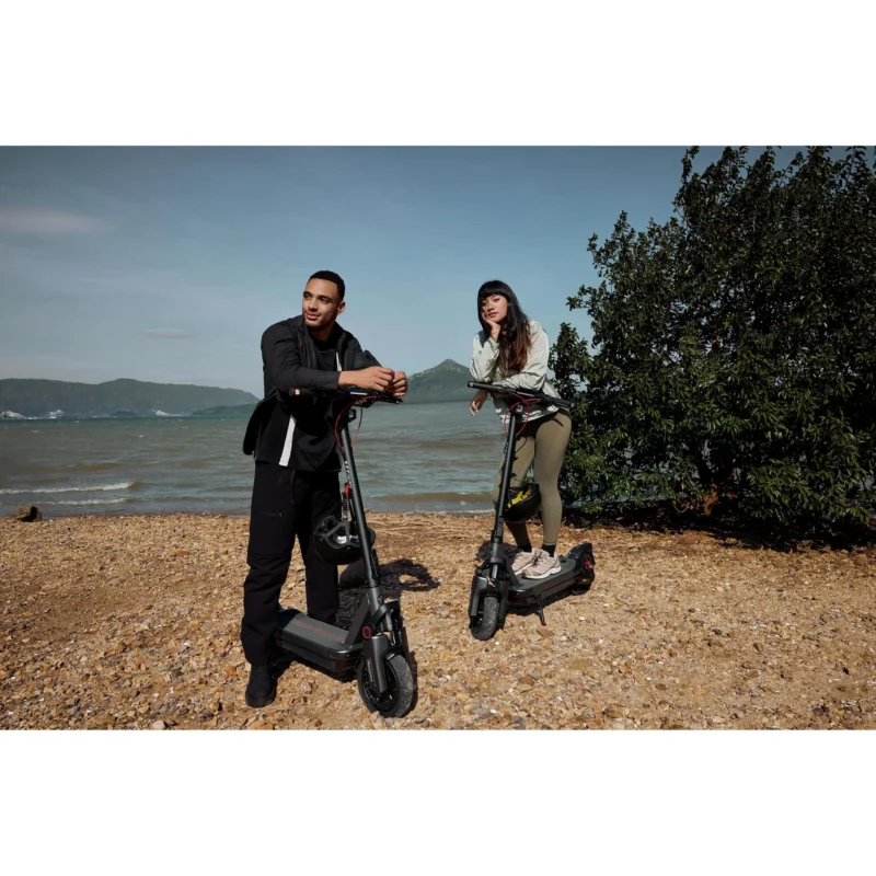 A young man and woman standing next to their Segway Ninebot KickScooter MAX G3 electric scooters on a pebble beach overlooking the sea.