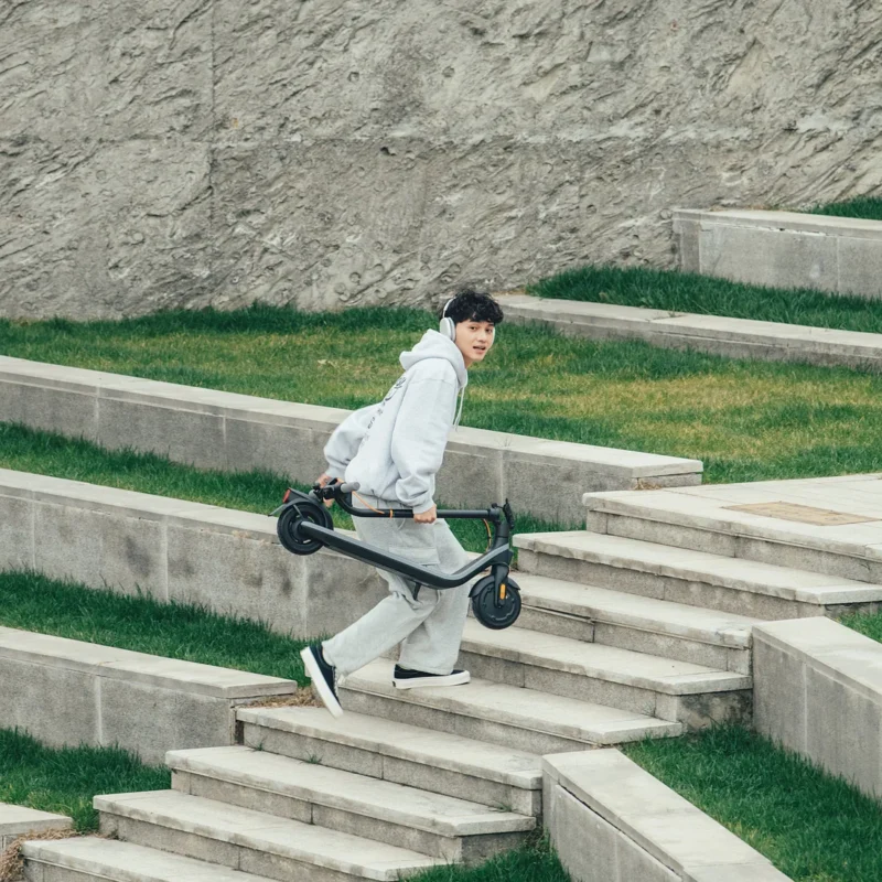 A young man wearing headphones carrying a folded Segway electric scooter as he walks up concrete outdoor stairs.