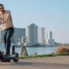 A man standing on an electric scooter by a waterfront path with a city skyline in the background.