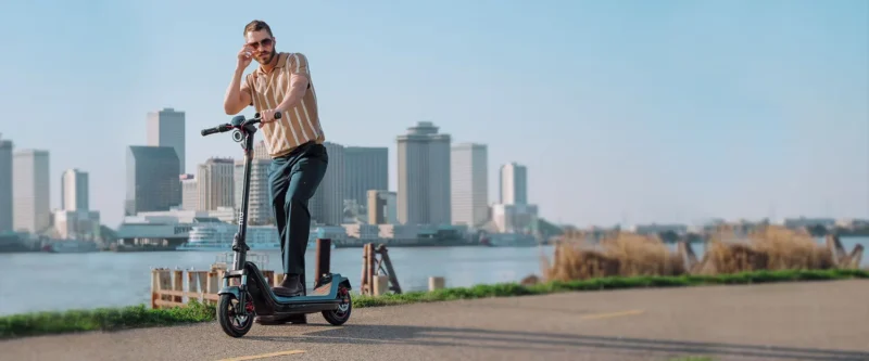 A man standing on an electric scooter by a waterfront path with a city skyline in the background.