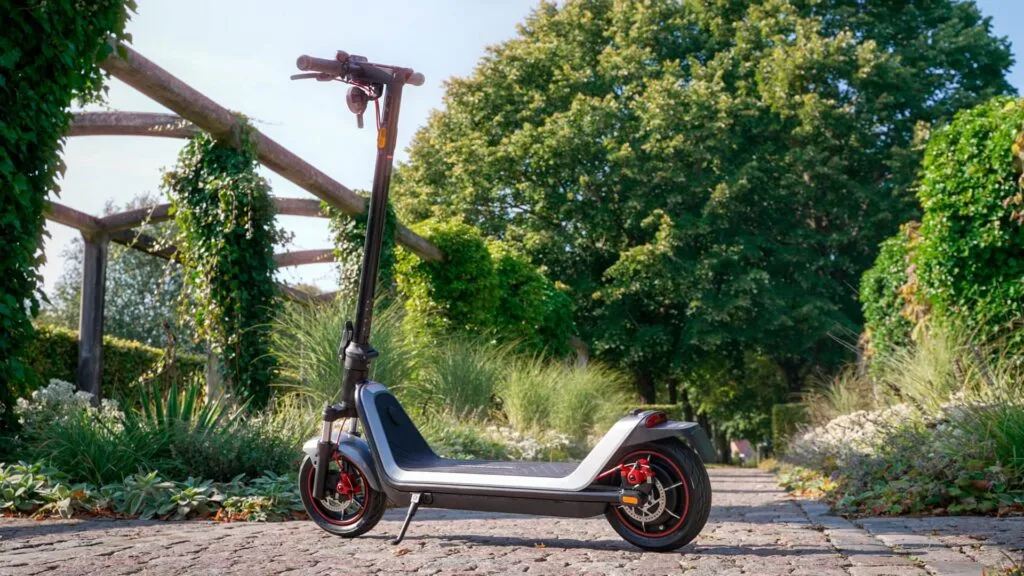 The NIU KQi300X electric scooter parked on a rustic cobblestone path surrounded by lush greenery and a wooden pergola structure.