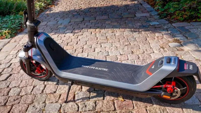 Close-up outdoor shot of the wide foot deck on the NIU KQi300X featuring the "MAKE LIFE ELECTRIC" branding text and kickstand down.