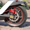 Close-up of the rear wheel hub motor, red brake caliper, and disc brake rotor of the NIU KQi300X scooter on a cobblestone path.