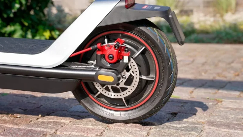 Close-up of the rear wheel hub motor, red brake caliper, and disc brake rotor of the NIU KQi300X scooter on a cobblestone path.