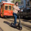 A man riding a NIU electric scooter down a city street alongside a historic red streetcar.
