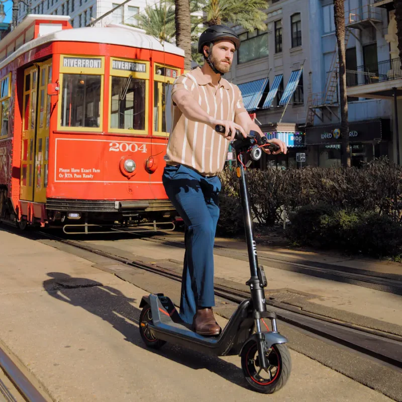 A man riding a NIU electric scooter down a city street alongside a historic red streetcar.