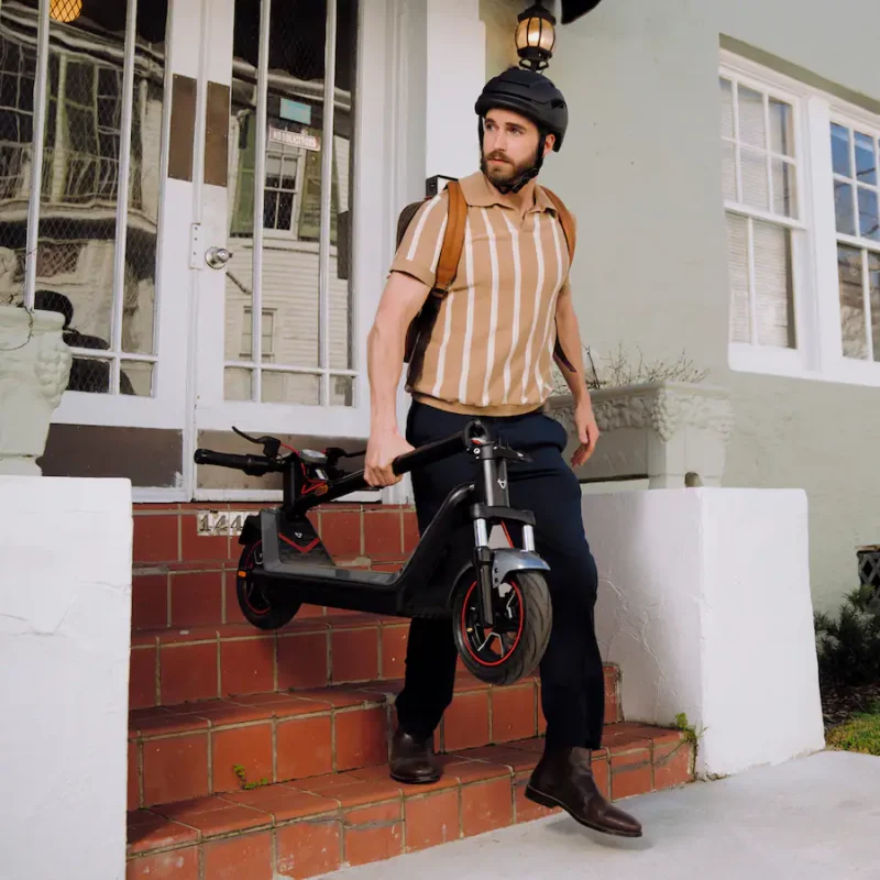 A man wearing a helmet and striped polo shirt carrying a folded electric scooter down house steps.