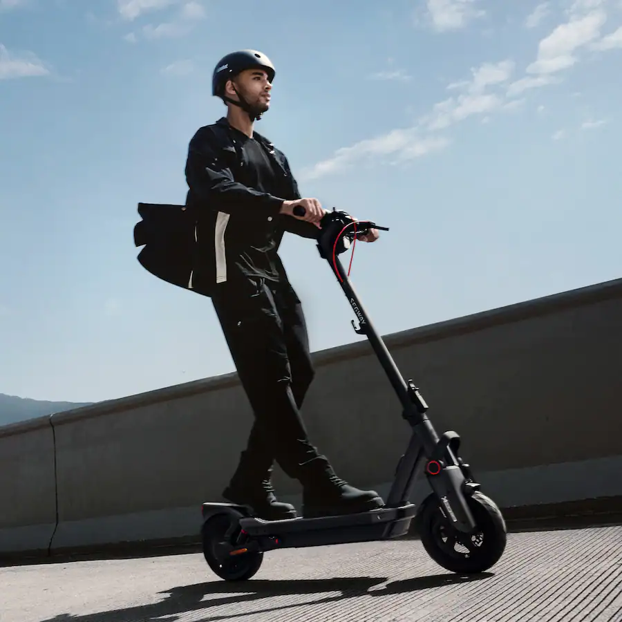 A young man wearing a helmet and messenger bag riding a Segway electric scooter along an elevated coastal pathway against a bright blue sky.