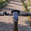 First-person rider perspective of the NIU KQi300X handlebars, central digital dashboard display, throttle, and bell over an outdoor path.