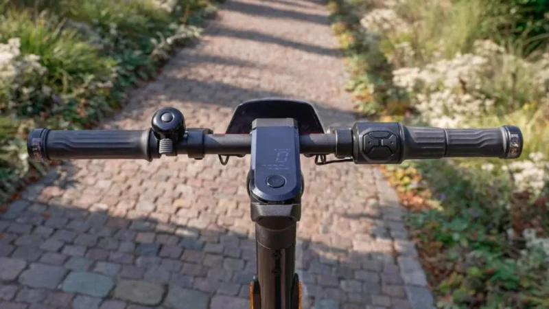 First-person rider perspective of the NIU KQi300X handlebars, central digital dashboard display, throttle, and bell over an outdoor path.
