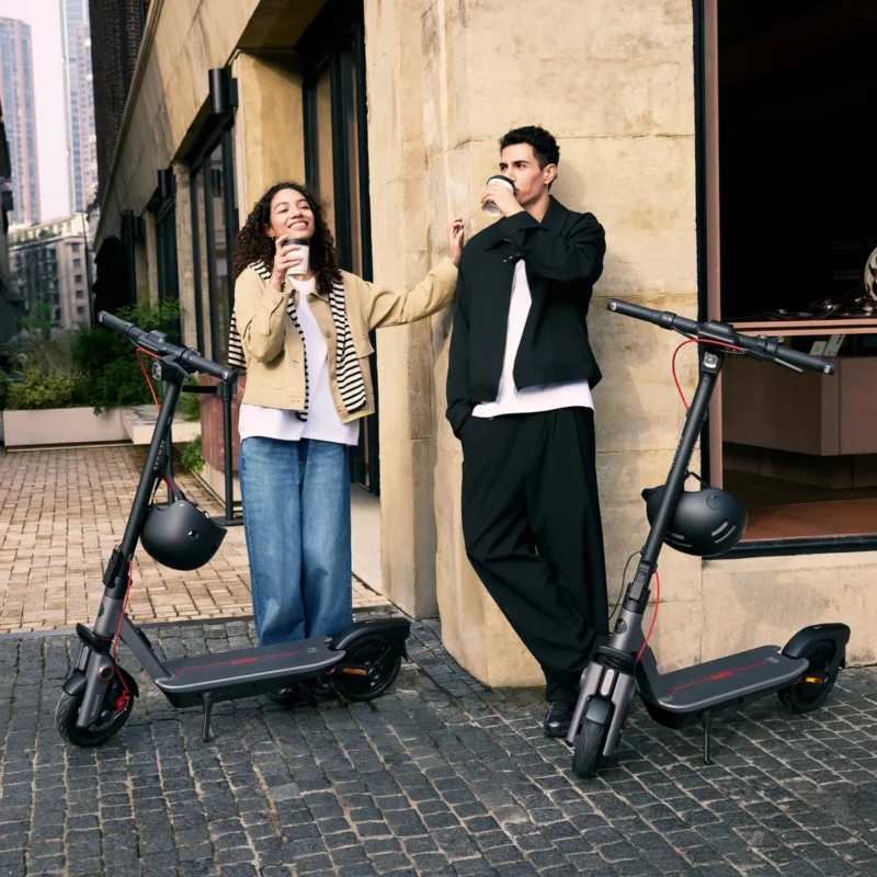 A smiling young woman and man holding coffee cups while leaning against a stone building next to two parked Segway electric scooters with mounted helmets.