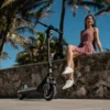 A woman in a floral dress and white sneakers is sitting on a low stone wall made of rough-cut blocks. The dark gray NIU KQi 200P electric scooter is parked next to her on a paved area. Palm trees are visible in the background against a clear blue sky.