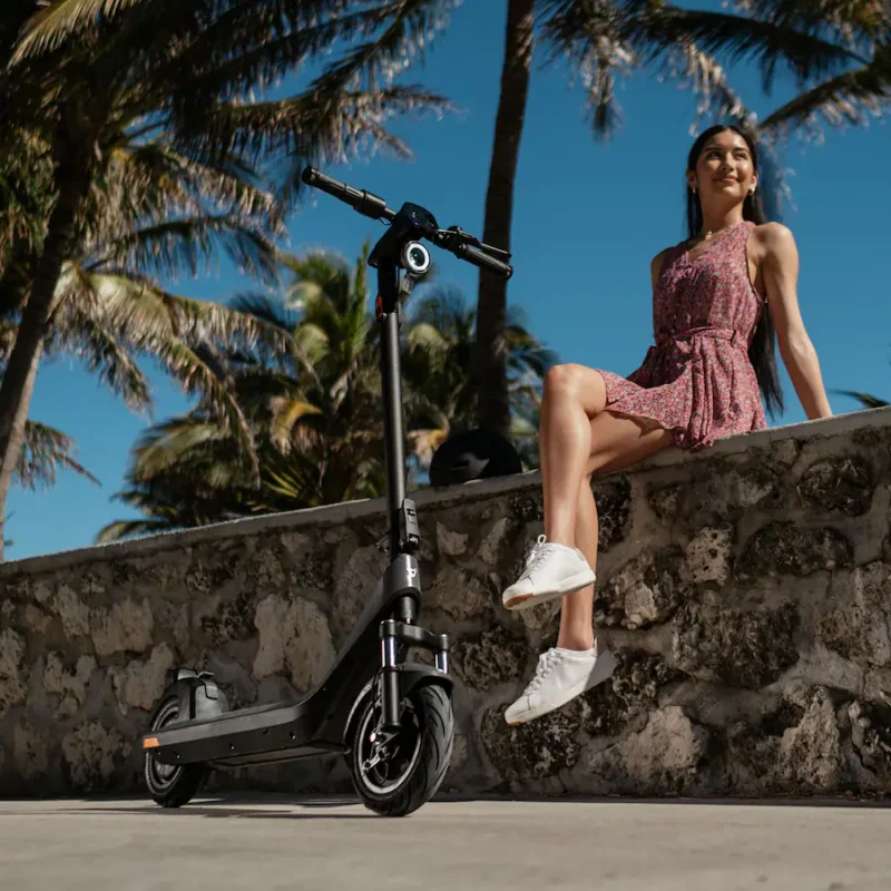 A woman in a floral dress and white sneakers is sitting on a low stone wall made of rough-cut blocks. The dark gray NIU KQi 200P electric scooter is parked next to her on a paved area. Palm trees are visible in the background against a clear blue sky.