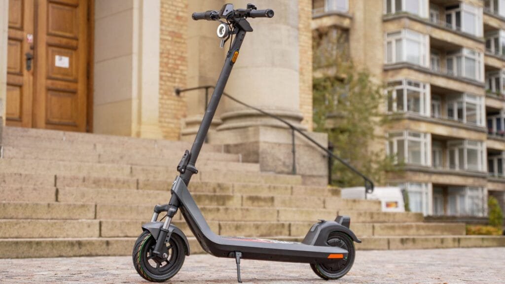 A three-quarter front view of the dark gray NIU KQi 200P electric scooter, parked on a brick pathway in front of large stone steps leading to a traditional building with wooden doors, under natural daylight.