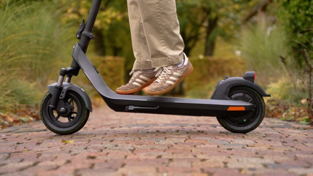 A close-up photograph capturing the lower half of a person wearing khaki pants and beige and brown sneakers standing on the dark gray deck of a NIU KQi 200P electric scooter, which is parked on a brick pathway in an outdoor setting with blurred greenery.