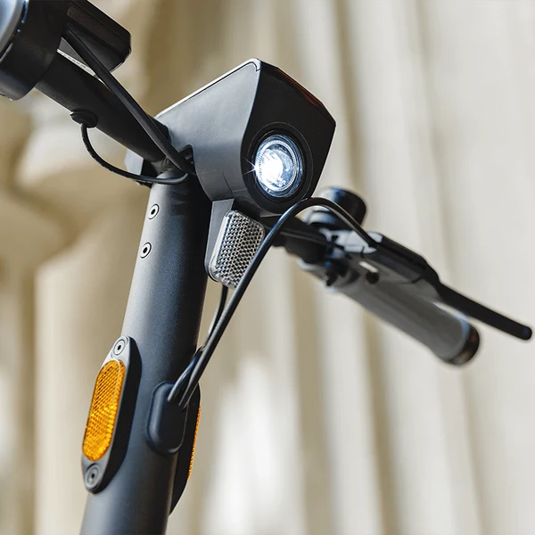 Close-up of electric scooter handlebars featuring a bright central LED headlight, reflector, and handlebar grips.