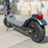 Rear wheel, tire tread, and illuminated red tail light of a gray electric scooter parked on a paved surface.