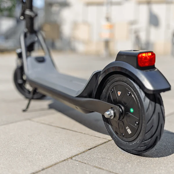 Rear wheel, tire tread, and illuminated red tail light of a gray electric scooter parked on a paved surface.