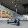 A dark gray electric scooter parked on its kickstand beneath an urban concrete overpass with colorful graffiti art.
