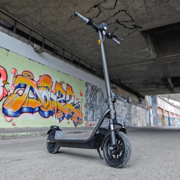 A dark gray electric scooter parked on its kickstand beneath an urban concrete overpass with colorful graffiti art.