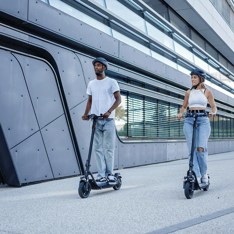 A man and a woman riding NIU KQi 100P electric scooters together on a paved city street past a modern building.