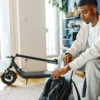 A young man sitting indoors packing a backpack next to a parked, folded NIU KQi 100P electric scooter.