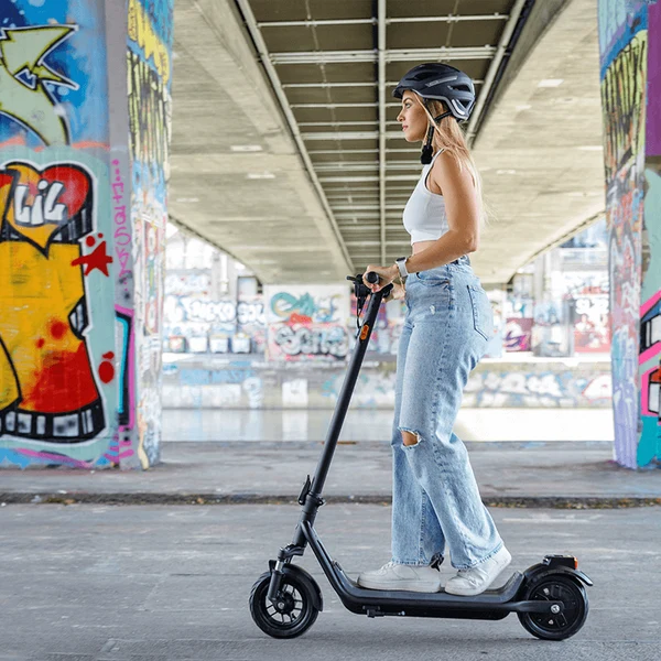 A woman wearing a helmet and casual clothing riding an electric scooter beneath a concrete bridge with graffiti art.