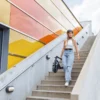 A woman wearing a helmet and casual clothes carrying a folded electric scooter down an outdoor concrete staircase.