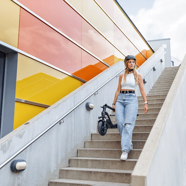 A woman wearing a helmet and casual clothes carrying a folded electric scooter down an outdoor concrete staircase.