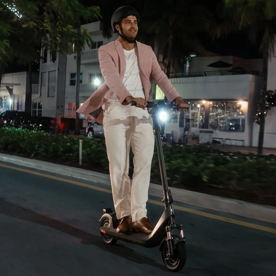 A man in a pink blazer and helmet is riding the dark gray NIU KQi 200P electric scooter down a street in Miami Beach at night. The background features blurred palm trees, neon lights, and Art Deco buildings. The scooter's headlight is on.