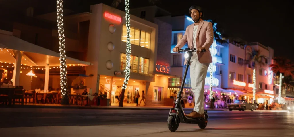 A man in a pink blazer and helmet is riding the dark gray NIU KQi 200P electric scooter down a palm-lined street in Miami at night. The street is illuminated by warm ambient lights from Art Deco buildings and neon signs, creating a vibrant city scene.