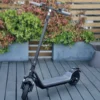 A modern electric scooter standing upright on a slatted wooden deck with outdoor planters and decorative pots in the background.