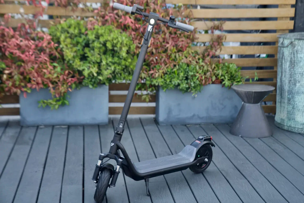 A modern electric scooter standing upright on a slatted wooden deck with outdoor planters and decorative pots in the background.