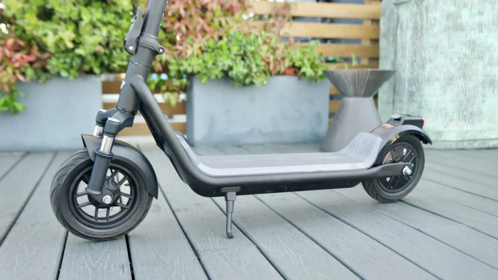 Full side profile of a dark gray electric scooter on a wooden deck, showing the dual tube front suspension and standing deck.