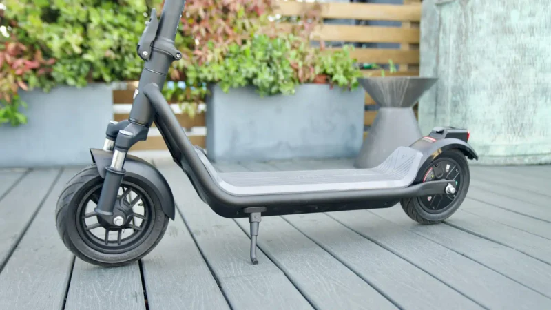 Full side profile of a dark gray electric scooter on a wooden deck, showing the dual tube front suspension and standing deck.