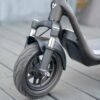 Close-up of the front wheel, tire, dual fork suspension, and side kickstand of a dark gray electric scooter resting on a wooden deck.