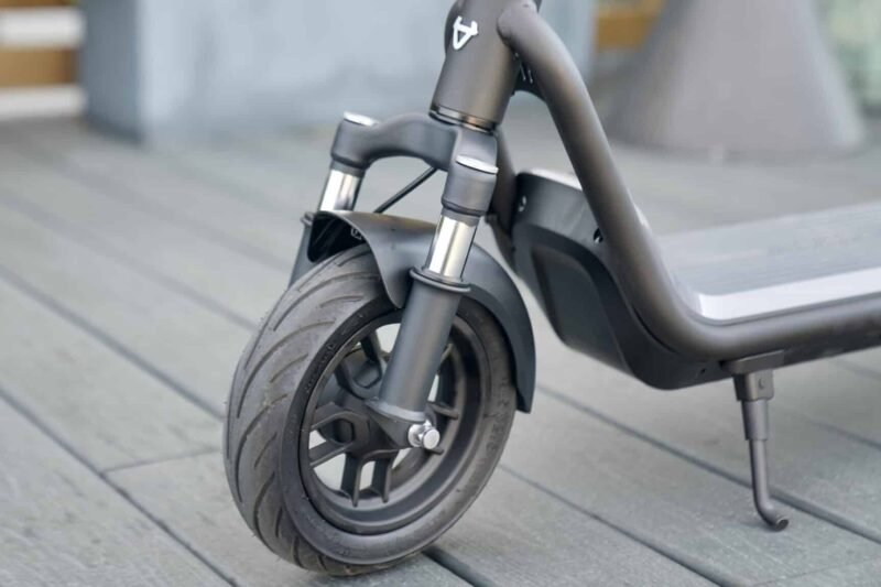 Close-up of the front wheel, tire, dual fork suspension, and side kickstand of a dark gray electric scooter resting on a wooden deck.