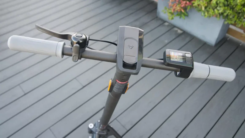 Overhead view of black electric scooter handlebars showing the brake lever, mechanical bell, center headlight module, and display.